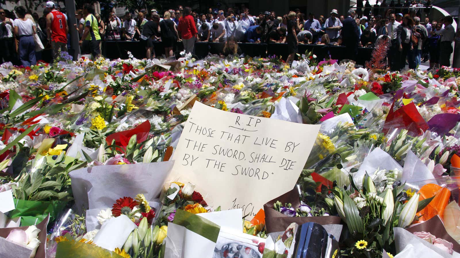 Members of the public stand behind a note that can be seen amongst floral tributes .