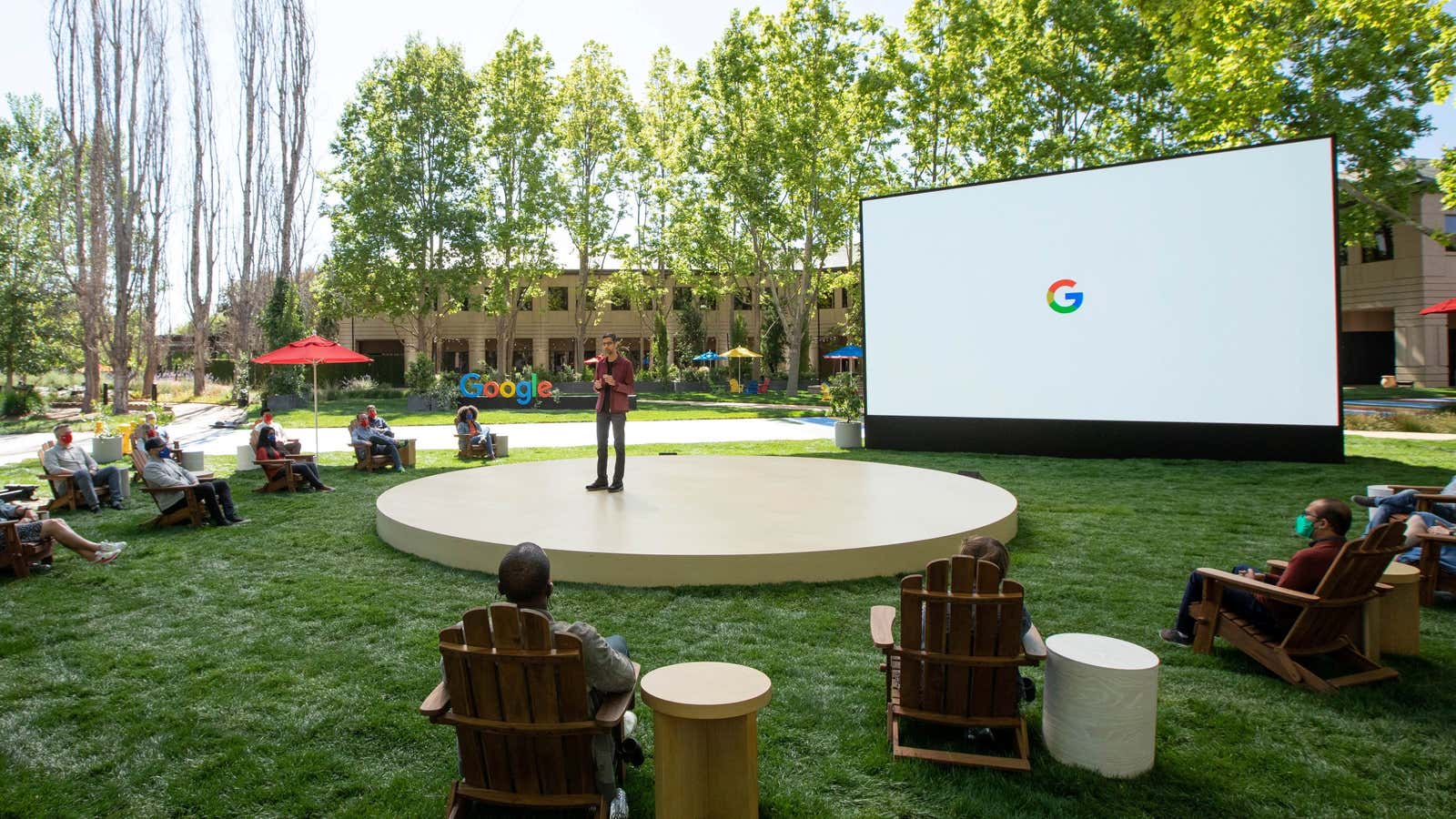 Google CEO Sundar Pichai stands on a circular outdoor stage ringed by attendees sitting in lawn chairs.