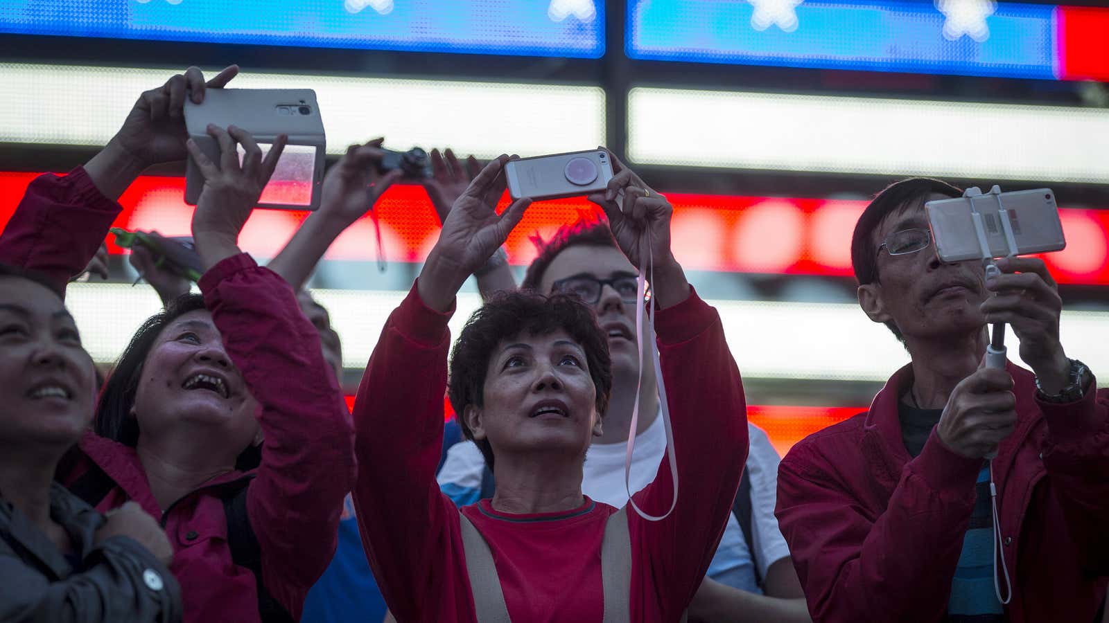 People hold up their mobile devices as they stand in front of a billboard owned by Revlon that takes their pictures and displays them at…