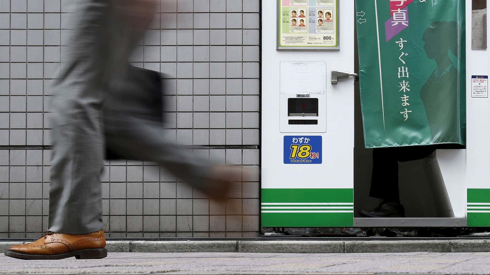 A pedestrian walks past an ID photo machine box as a man sits inside at a business district in Tokyo, September 15, 2015. The Bank of Japan said on Tuesday that slowing emerging market demand was putting further strains on the economy but held off on expanding stimulus, preserving its limited policy options in case an expected U.S. rate hike sparks more global volatility. REUTERS/Yuya Shino – GF10000206357