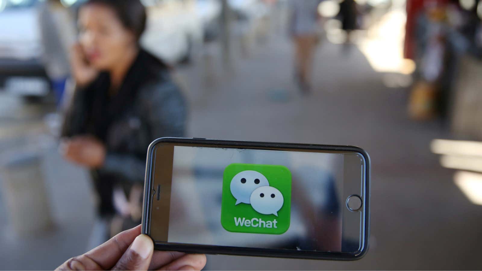 WeChat logo is displayed on a mobile phone as a woman walks past as she talks on her mobile phone at a taxi rank in…