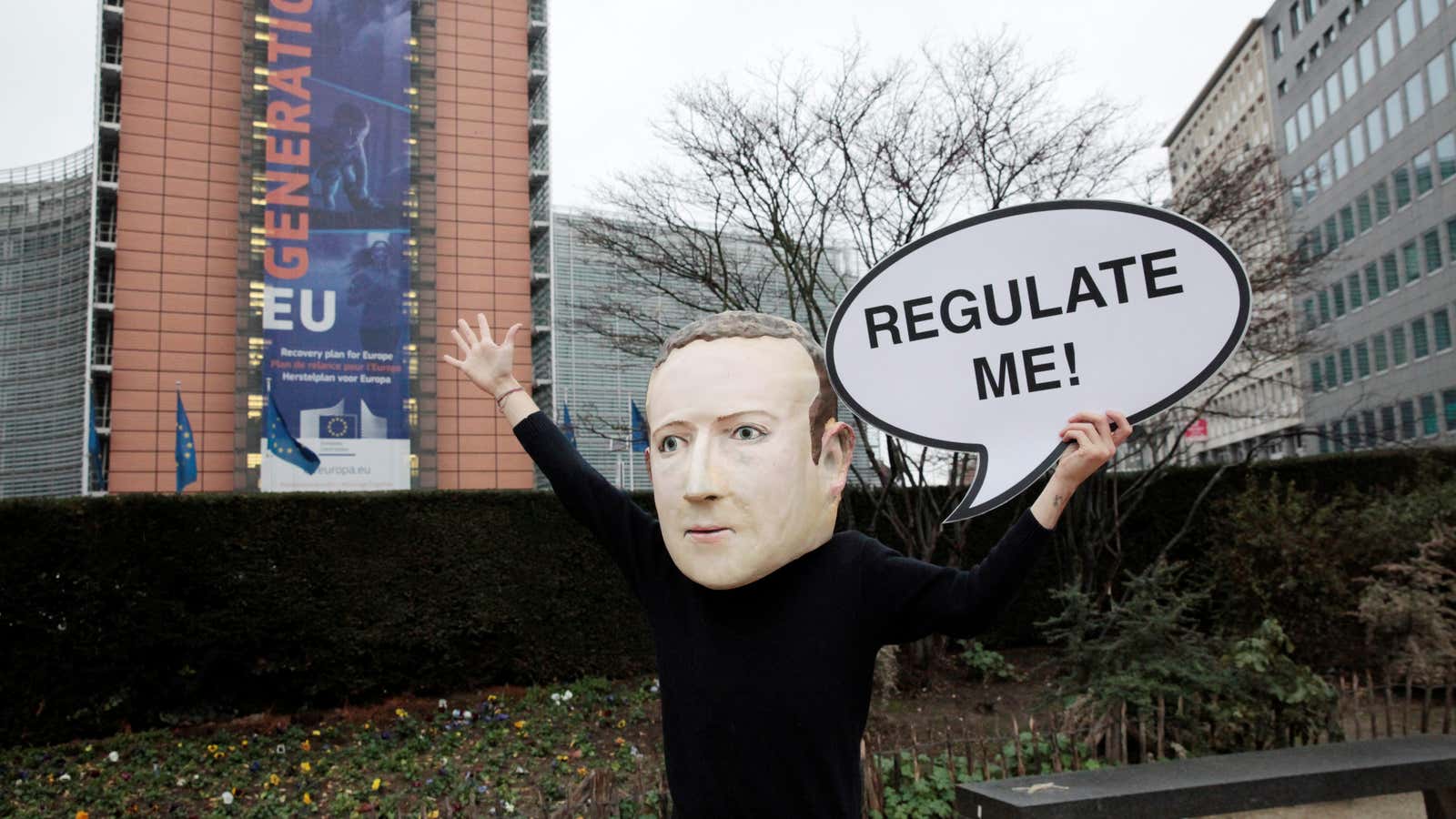 An activist wearing a paper mache Mark Zuckerberg mask holds a sign that says “Regulate me!”