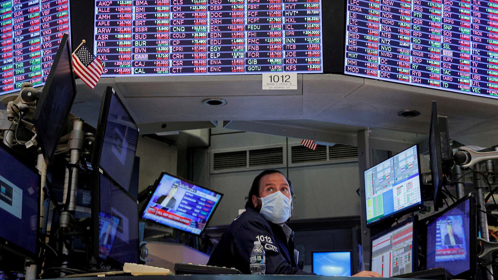 A trader wearing a face mask works inside a booth at the New York Stock Exchange beneath a screen showing stock prices.