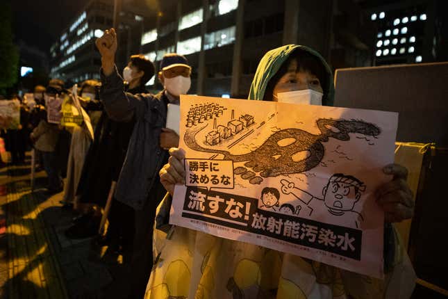 People demonstrate outside of the prime minister's official residence in Tokyo Japan. A woman wearing a white mask holds up a poster with a drawing depicting wastewater spilling from the Fukushima plant into the ocean and creating the shape of a skull.