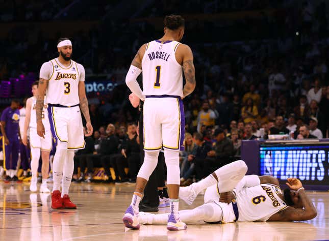 During an NBA basketball game, a Black man in a white uniform writhes on the wood floor in pain, grabbing his testicles, while his teammates look on.