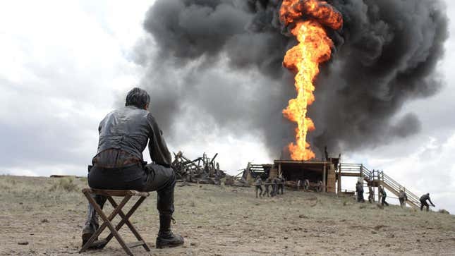 A screenshot from There Will Be Blood of a man in early 20th century period dress (Daniel Day Lewis) seated facing away from us on a dusty outdoor vista, with a burning oil derrick visible in the background