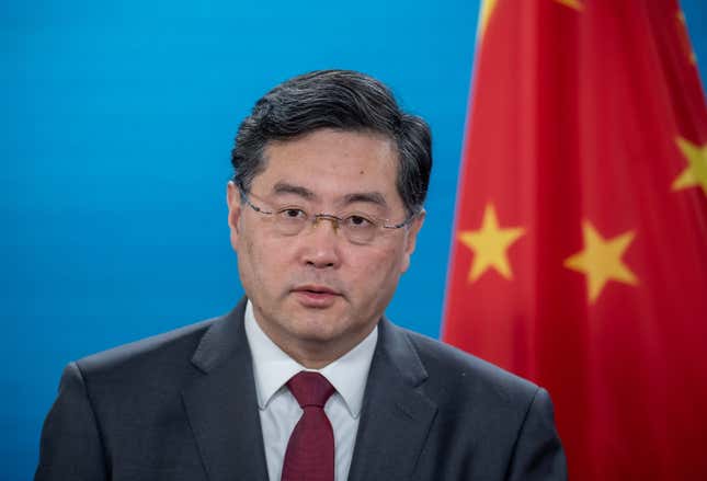 A portrait of Qin Gang wearing a gray suit and maroon tie, standing in front of a blue background and a Chinese flag.