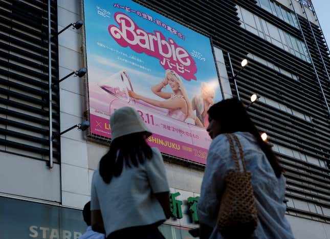 Two girls stand in front of a Barbie poster on the side of a building in Japan.