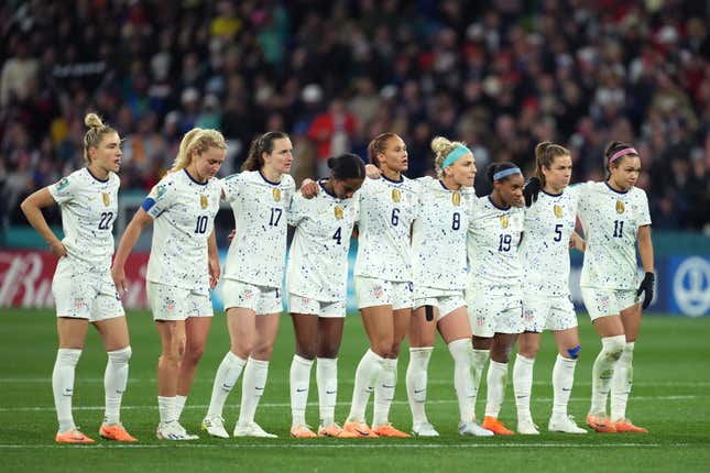 A group of women, all with their hair up, wear matching white soccer uniforms and stand with their arms around each other, looking towards the goal.