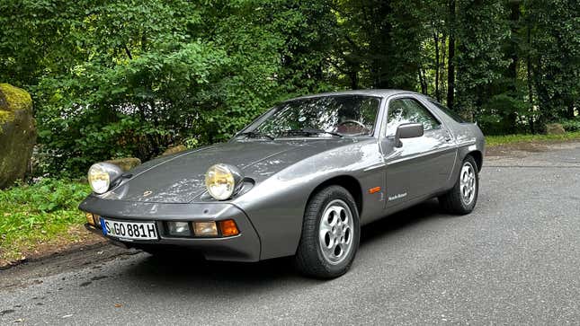 A dark gray 1981 Porsche 928 S 50 Jahre is parked in the Black Forest in Germany. Its headlights are up.