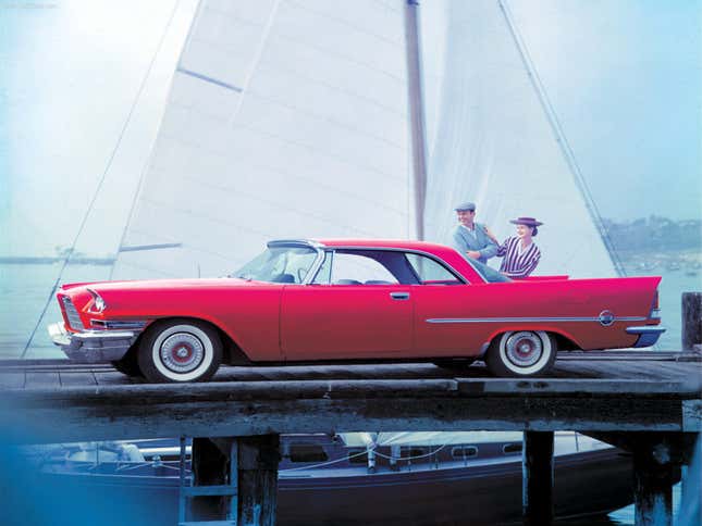 A red 1957 Chrysler 300C is parked on a dock in front of a sailboat