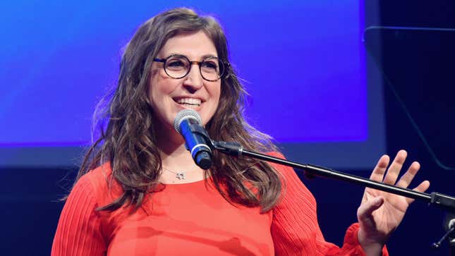 Mayim Bialik, a woman wearing glasses and an orange top, smiles behind a microphone.