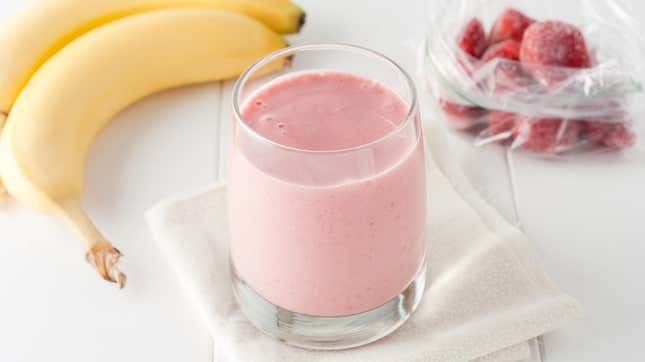 Strawberry smoothies in a clear glass sits on a white napkin with a bag of frozen strawberries and two bananas in the background