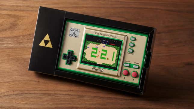 The gold and green Zelda 35th anniversary Game & Watch sitting in a black box emblazoned with a triforce symbol.