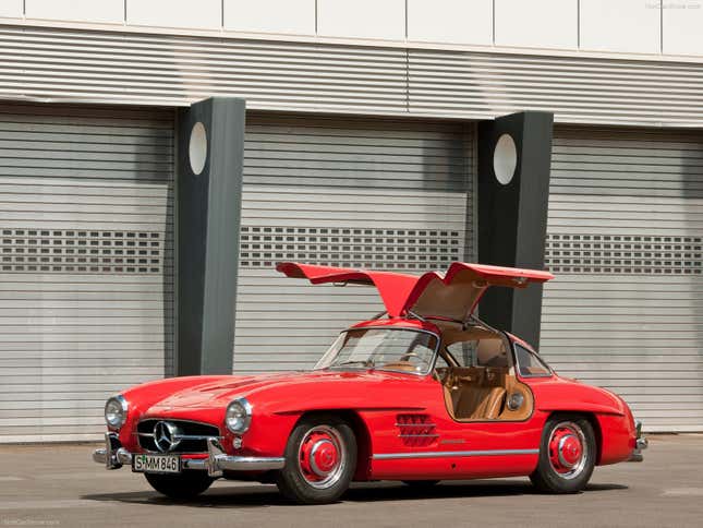 A red 300SL gullwing is parked with its doors up in front of some garages