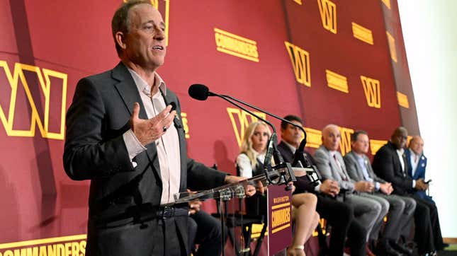 A slight white man in a suit without a tie stands at a podium in front of a maroon and gold background while six people sitting next to him in chairs look on.