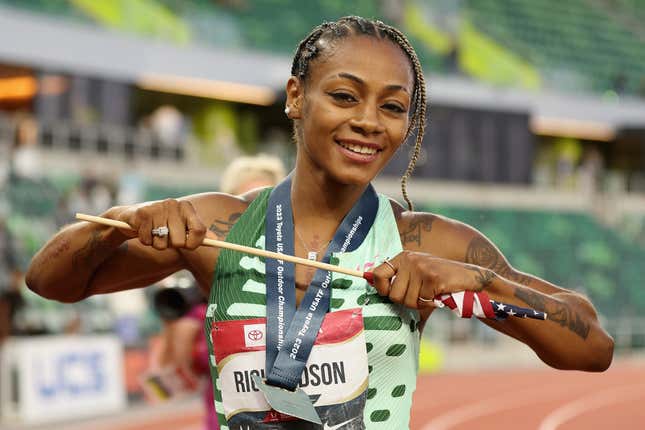 A smiling Black woman with her hair in braids and a green track uniform holds an Americn flag across her chest and sports a medal around her neck.