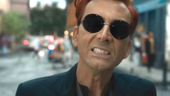 A screenshot of David Tennant in Good Omens Season 2 standing on the street wearing a black suit and dark sunglasses and grinning maniacally 