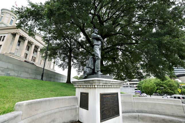A statue of a soldier on a white plinth takes up the middle of the photo. Behind the statue is a tree, and to the left is a greco-roman style building.