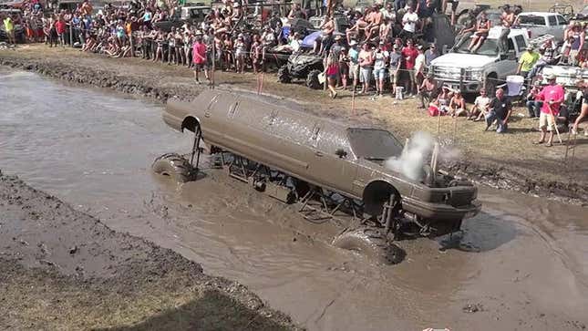 A mud-covered monster truck-like limousine wades through an artificial bog