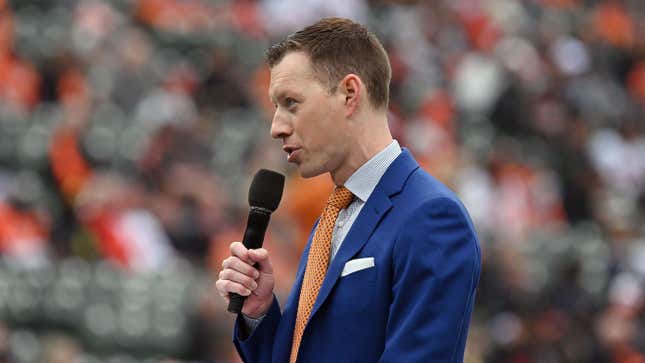 A young white man in a blue suit and orange tie, holds a microphone and talks to the crowd at Camden Yards before the Orioles home opener.