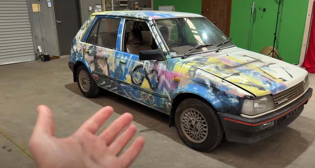 A spraypainted Daihatsu Charade is being gestured at while in a garage.