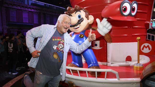 Voice actor Charles Martinet is seen smiling and giving a thumbs-up near a statue of Mario promoting Super Mario Odyssey.