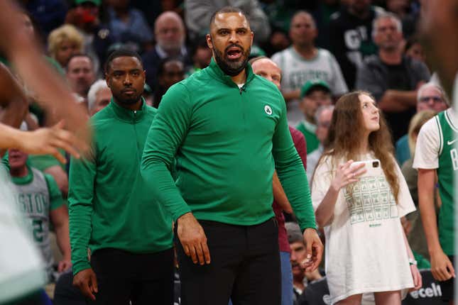 A Black man in a kelly green Celtics pullover and black pants stands on the sidelines of an NBA game, coaching the players on the floor.