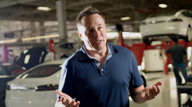 Elon Musk stands in front of several Tesla Model S vehicles on lifts. He is speaking to reporters while employees work on the cars in the background.
