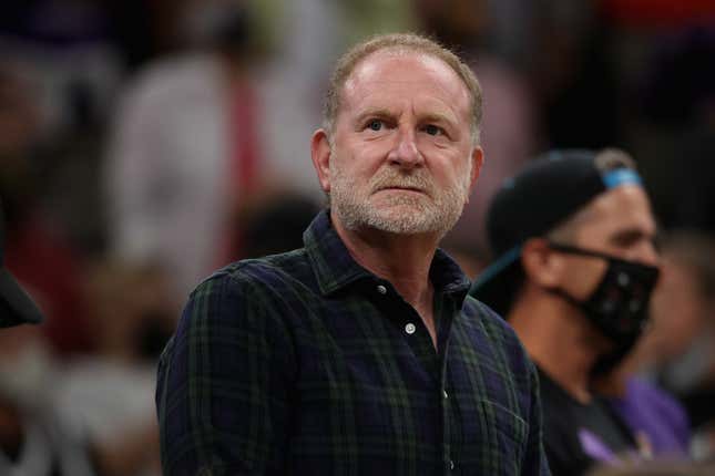 A middle-aged white man with a gray beard and short blonde hair wears a blue and green plaid shit at an NBA basketball game.