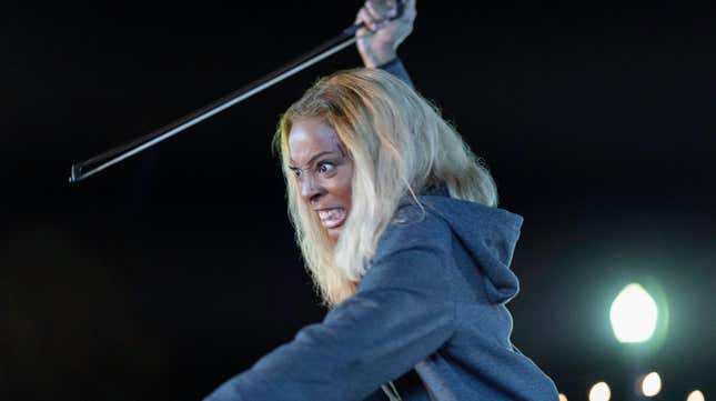 A woman wearing a blue hoodie raises a violin bow as a weapon with a crazed look on her face.