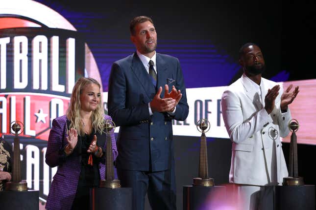 A white woman with long blonde hair in a purple and black suit, a tall white man with blonde hair and a navy blue suit, and a Black man with a beard and white suit applaud on the stage of the 2023 Naismith Basketball Hall of Fram Induction ceremony.