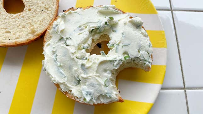 A halved bagel on a plate, covered in cream cheese and chives spread, with a bite taken out of it.
