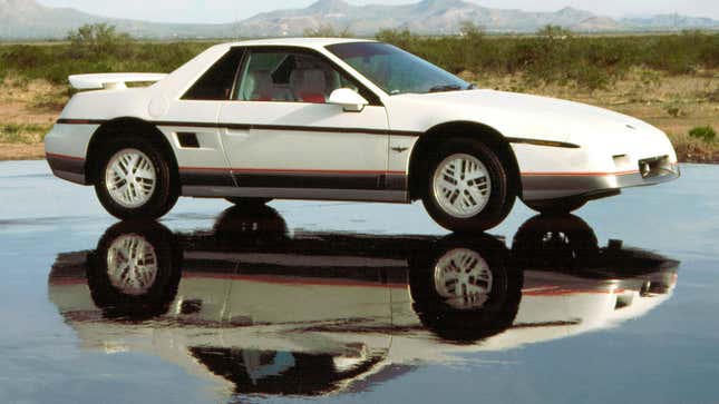 A photo of a white Pontiac Fiero sports car parked next to a puddle. 