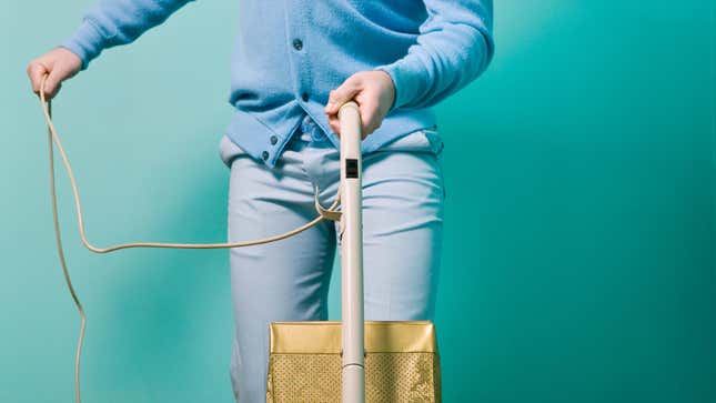 Head-on photo of a person wearing a light blue cardigan and gray jeans operating a vintage bagged vacuum with their left hand and holding the cord with their right. They're standing in front of a turquoise backdrop.