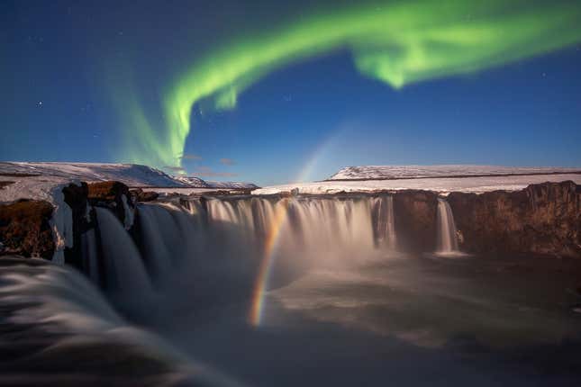 The northern lights arc above a waterfall creating a rainbow.