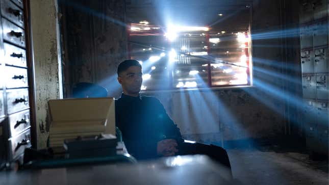 Gravik, in human form, sits at a desk, light streaming through the window behind him. 