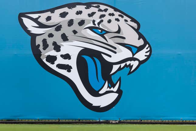 The Jacksonville Jaguars logo, a yellow jaguar head on a teal background, hangs from a fence at and NFL football game.