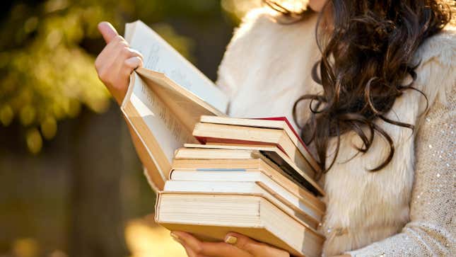woman holding a stack of books in one arm and an open book she's reading in the other hand