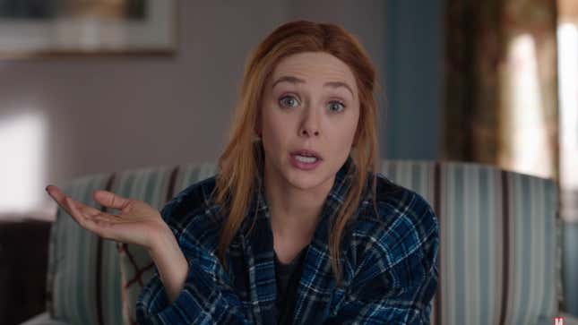 Wanda Maximoff, in a blue plaid shirt, sits on a striped couch and talks directly into the camera.