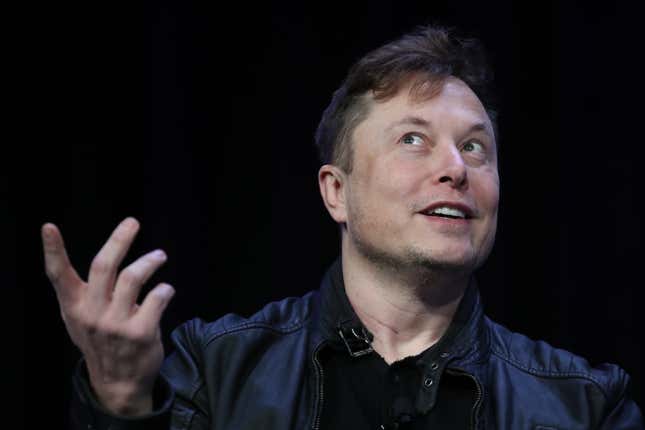 Elon Musk smiles and points up, talking at a conference in front of a black background