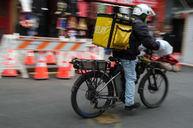 A delivery worker rides an electric bicycle through the streets of Manhattan, wearing a bright yellow "Relay" backpack.