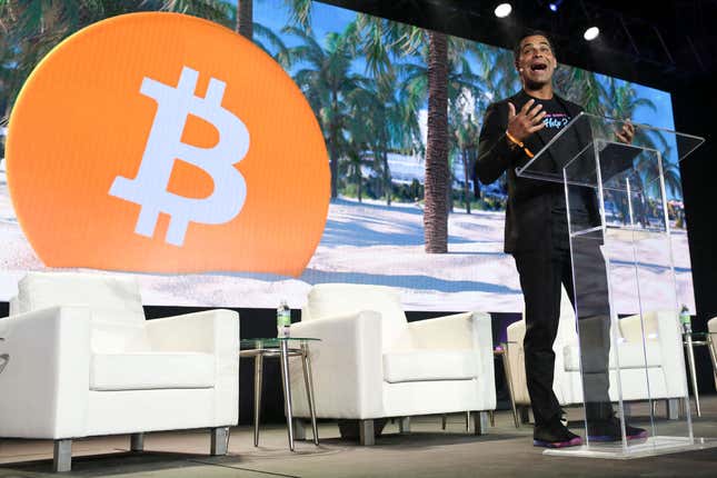 A person standing at a clear podium speaks on a stage. Behind the person are three white chairs and a backdrop of a tropical landscape with a large orange circle with the bitcoin symbol inside of it.
