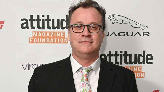 Russell T. Davies, in a black jacket and floral tie, stands before a placard reading attitude Magazine Foundation.