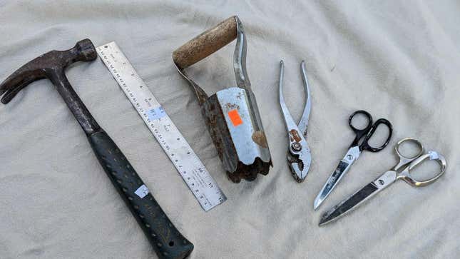 Estate sale tools laid out on a canvas tarp: A hammer, a ruler, a hole digger, a pair or pliers, and two pairs of heavy duty scissors