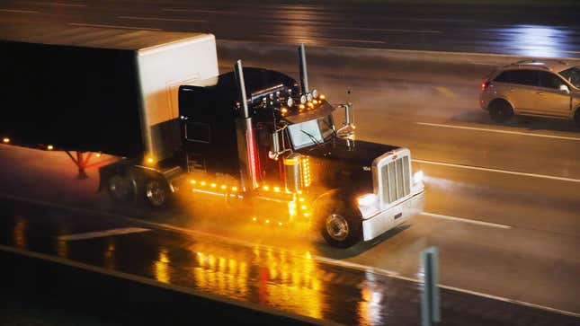 A photo of a big American truck driving in the rain, at night. 