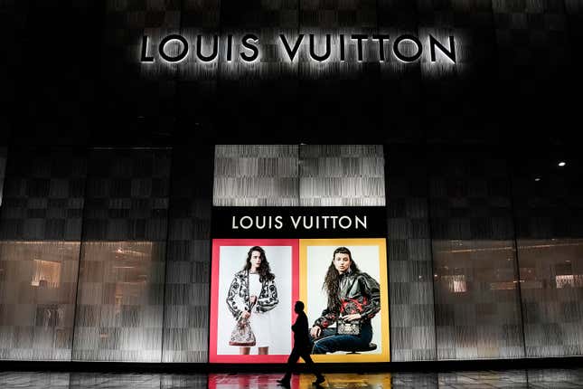 A pedestrian in silhouette walks in front of the illuminated billboards of a Louis Vuitton store at night.