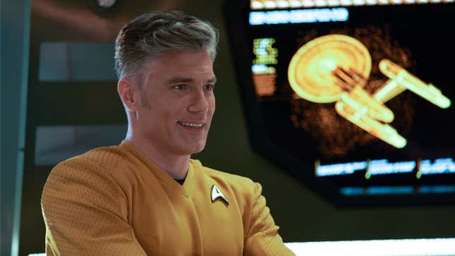 Pike, in his orange captain's uniform, smiles in front of a computer display of the Enterprise.