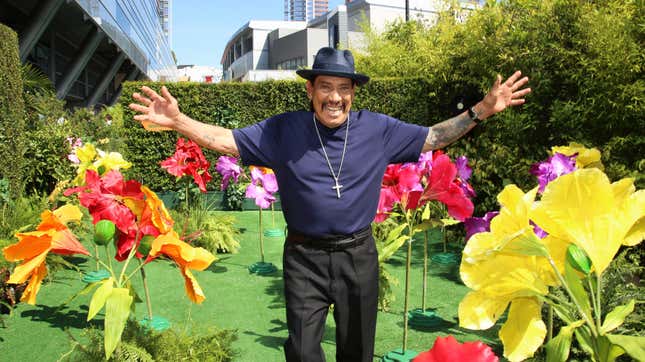 Actor Danny Trejo smiles and holds his arms out in a bright field of flowers.