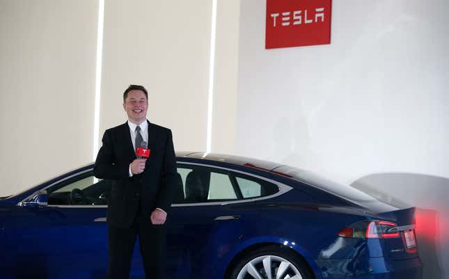 Elon Musk stands in front of a Blue Tesla Model S with a microphone in his hand. The red Tesla logo is on a white blank wall behind him.